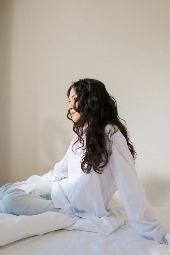 Young Woman with Black Curly Hair Sitting in Bed in White Pyjamas